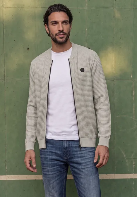 Homme en gilet clair et jeans, debout de manière décontractée devant un mur vert.