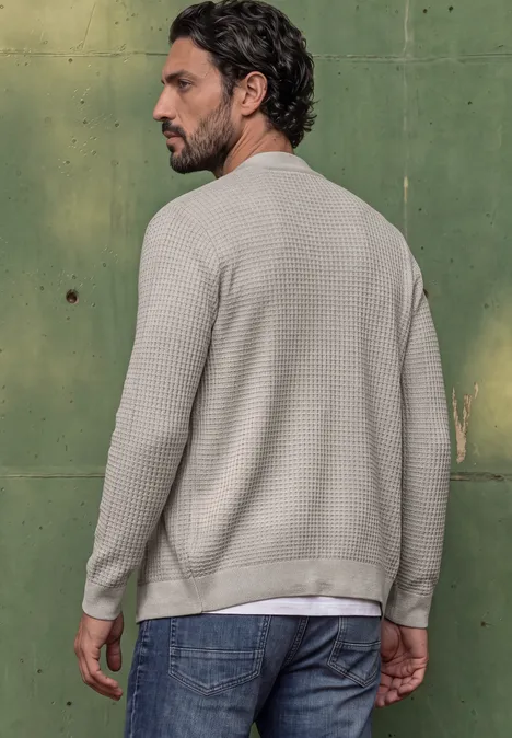 Un homme de profil porte un t-shirt beige à texture gaufrée et un jean devant un mur métallique vert.