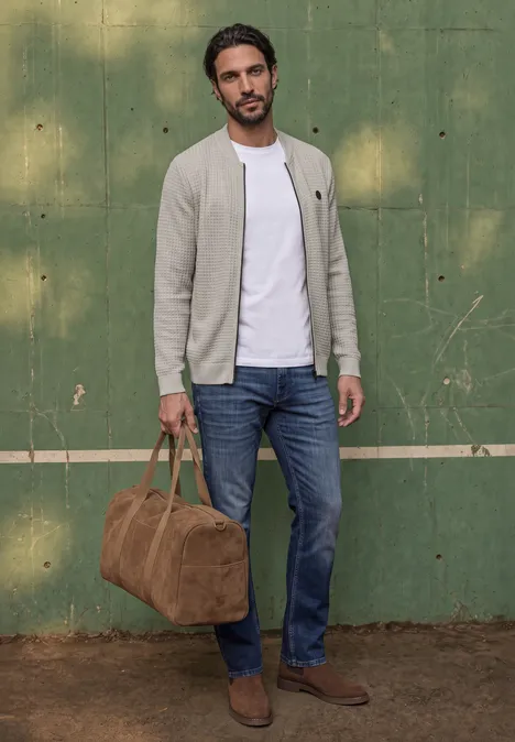 Un homme en jean, t-shirt blanc et veste grise, bottines et sac de voyage en daim marron devant un mur vert.