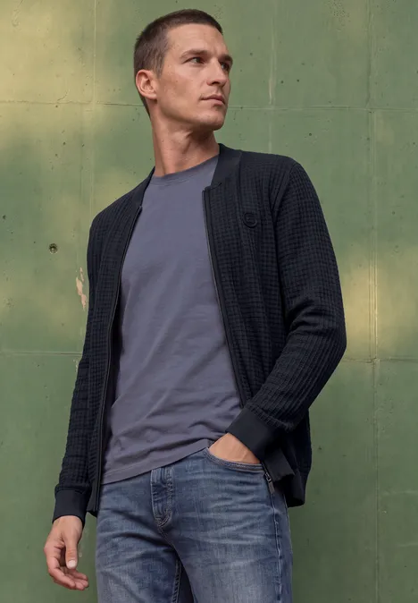 Un homme devant un mur vert, portant un t-shirt gris, un gilet en maille et un jean.