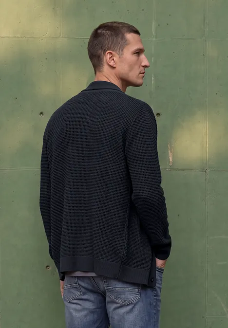 Un homme en veste sombre et jean de profil devant un mur vert.