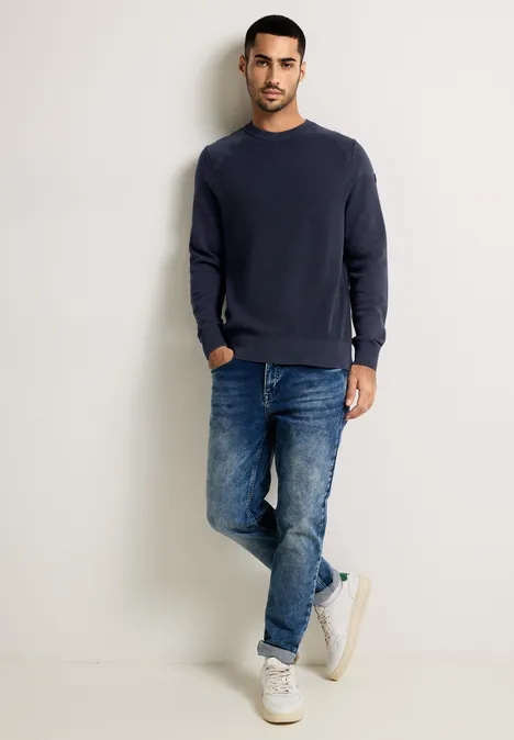 Homme en pull bleu foncé, jean et baskets blanches devant un mur clair.