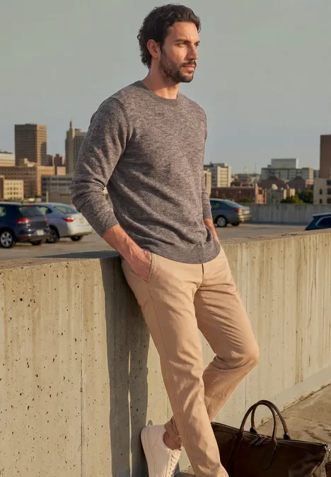 Ein Mann lehnt entspannt an einer Betonmauer, trägt Pullover und Chinohose, mit Tasche am Boden, Stadt im Hintergrund.