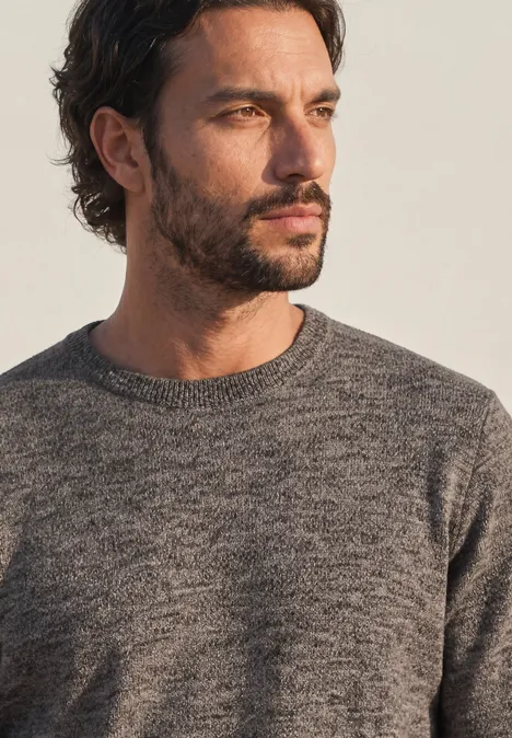 Ein Mann mit dunklen Haaren und kurzem Bart trägt einen grauen Strickpullover und blickt nach rechts.
