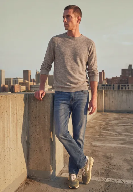 Ein junger Mann lehnt lässig an einer Betonmauer auf einem Parkhausdach, mit Skyline im Hintergrund.