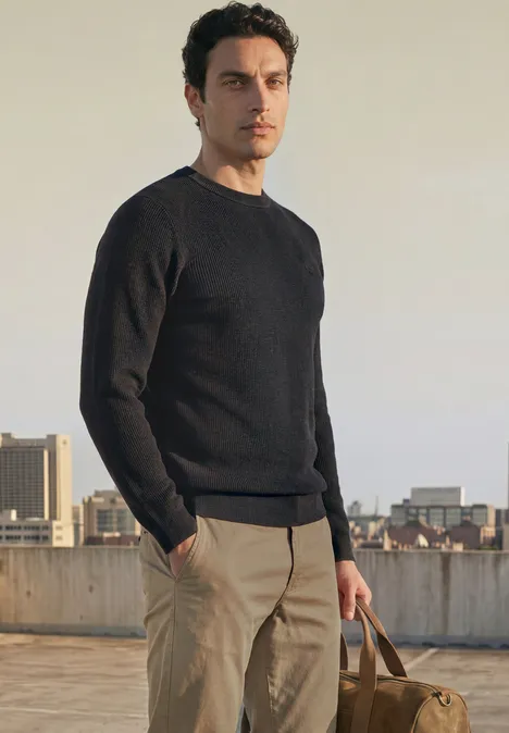 Trui met ronde hals en gemengde texturen Black Een man in een zwarte trui en lichte broek staat op een dak en houdt een bruine tas in zijn hand.