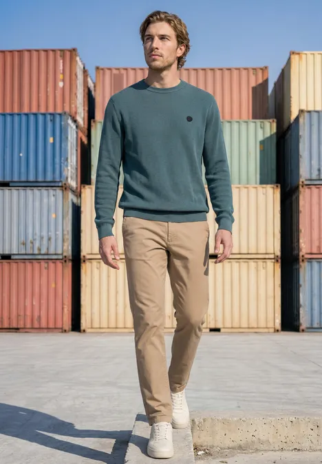 Trui met ronde hals en gemengde texturen cold blue Een man in een groene trui en beige broek loopt langs gekleurde containers in een industriële haven.