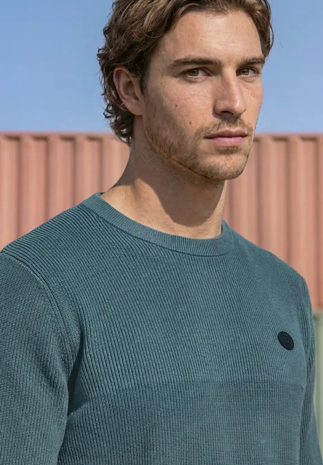 Trui met ronde hals en gemengde texturen cold blue Portret van een jonge man in een turquoise trui voor een roze container, peinzende blik.
