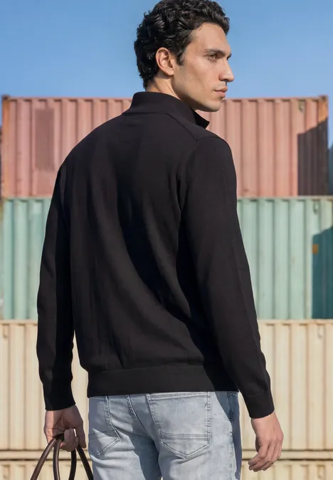 Basic Pullover mit Troyerkragen Black Ein Mann steht in schwarzem Pullover und hellen Jeans vor bunten Containern und hält eine Tasche.
