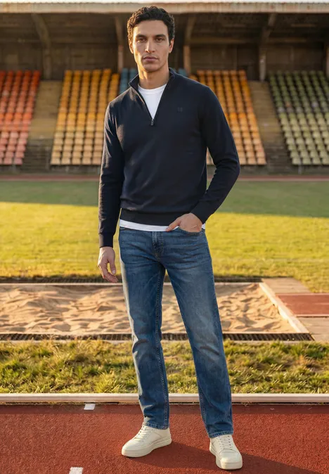 Basic Pullover mit Troyerkragen dark basic blue Ein junger Mann in Jeans, Sneakern und Pullover steht selbstbewusst auf einer Laufbahn vor Tribünen.
