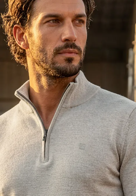 Ein Mann mit Bart und dunklen Locken trägt einen hellgrauen Pullover mit Teil-Reißverschluss und schaut zur Seite.
