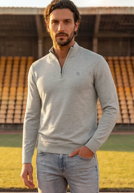 Ein Mann in grauem Pullover und Jeans steht in einem Stadion mit leeren Tribünen im Hintergrund.