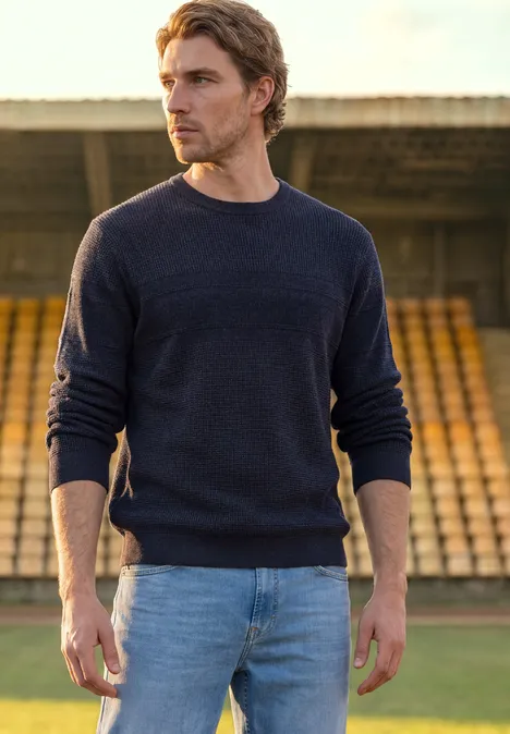 Pullover mit Rundhals und Struktur dark basic blue melange Ein Mann in dunklem Pullover und Jeans steht vor leeren Tribünen im Freien.