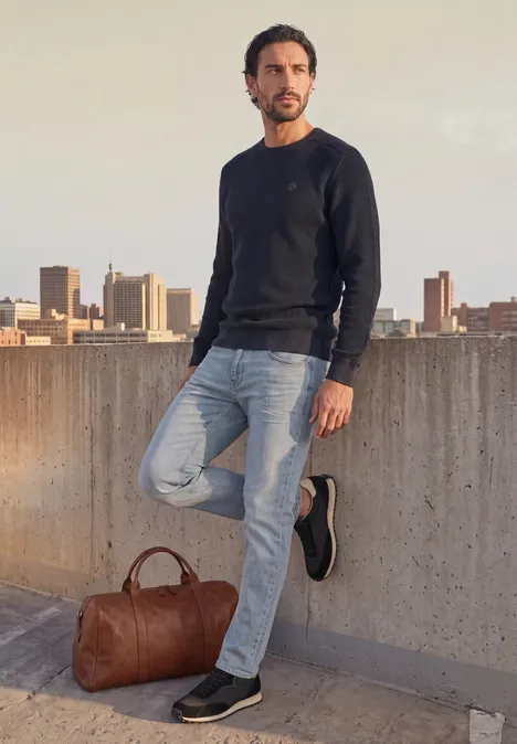 Trui met ronde hals en schouderdetail dark basic blue Man in een casual trui en spijkerbroek leunt op een dakterras met stadsgezicht op de achtergrond.