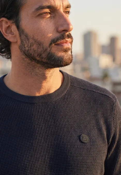 Trui met ronde hals en schouderdetail dark basic blue Een bedachtzame man met baard en donker trui voor een vage stadsgezicht in avondlicht.