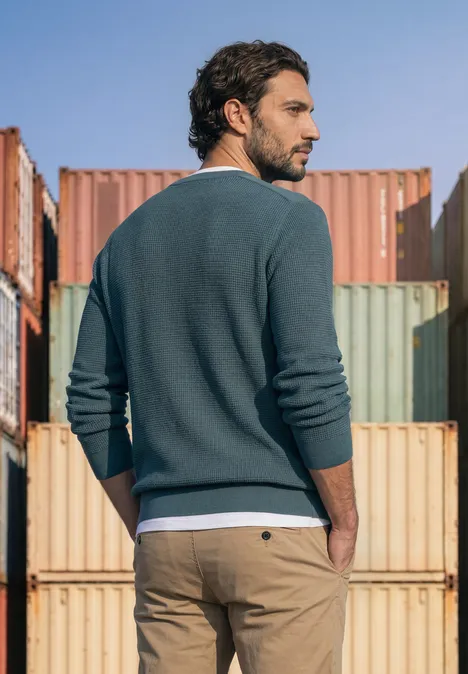 Trui met ronde hals en schouderdetail cold blue Een man in trui en chino staat voor kleurrijke containers en kijkt opzij.