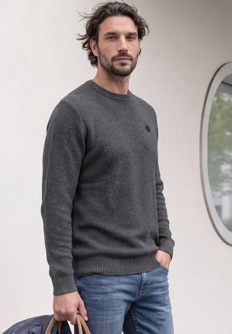 Ein Mann in grauem Pullover und Jeans steht vor einer Wand mit rundem Fenster.
