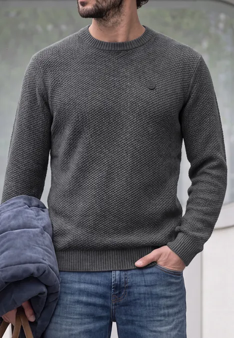 Ein Mann trägt einen grauen Strickpullover und Jeans, hält eine Jacke in der Hand.