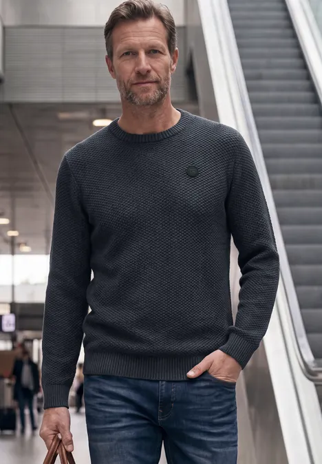 Ein Mann in Jeans und Pullover steht in einer öffentlichen Umgebung vor einer Rolltreppe.