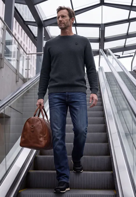 Ein Mann in Jeans und Pullover hält eine braune Ledertasche auf einer Rolltreppe in einem modernen, verglasten Gebäude.