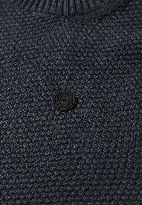 Nahaufnahme eines dunkelblauen, strukturierten Strickpullovers mit rundem, schwarzem Logo-Button in der Mitte.
