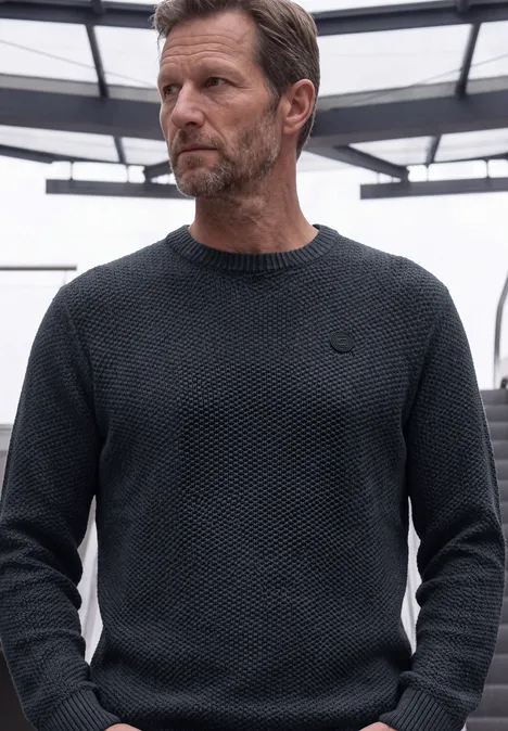 Ein Mann trägt einen dunkel gestrickten Pullover und blickt nach links vor einer modernen Kulisse.
