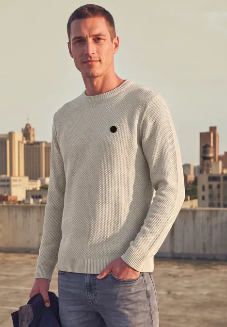 Ein Mann in einem hellen Pullover steht auf einem Dach, im Hintergrund die Stadt.