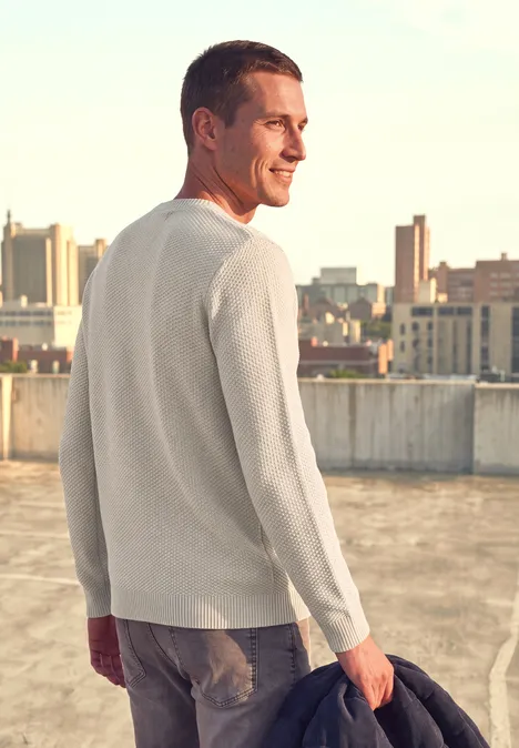 Ein Mann in hellem Pullover hält eine Jacke und schaut auf eine Stadtlandschaft vom Dach aus.