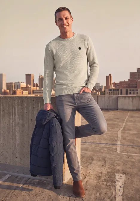 Ein Mann lehnt entspannt auf einem Parkdeck, trägt lässigen Pullover, Jeans und hält eine Jacke.