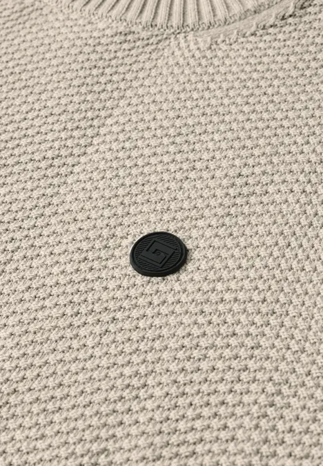 Beiger Pullover mit Waffelstrickmuster und einem kleinen, runden schwarzen Markenlogo in der Mitte.