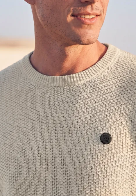 Nahaufnahme eines Mannes im cremefarbenen Strickpullover mit rundem Emblem auf der Brust.