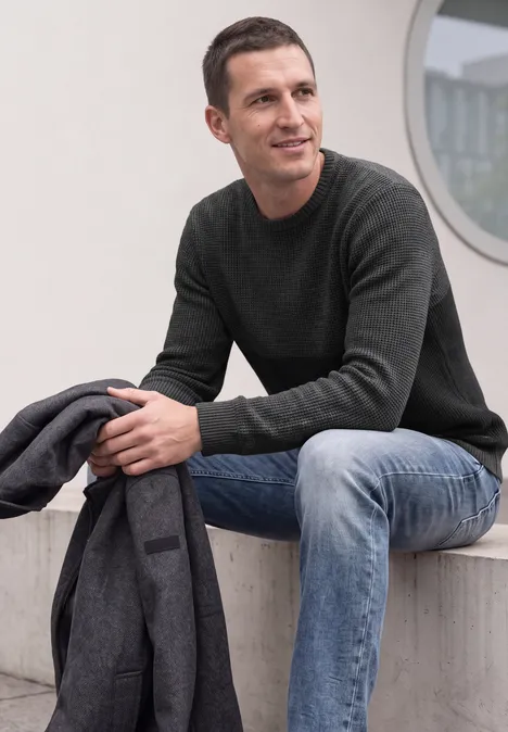Ein Mann sitzt vor einem Gebäude, trägt Jeans und Pullover, hält eine Jacke und blickt seitlich.