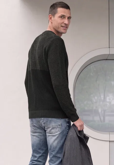 Ein Mann schaut über die Schulter, trägt Jeans, dunklen Pullover und hält eine graue Jacke.