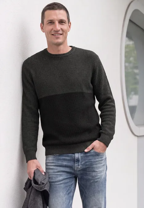 Ein Mann in Pullover und Jeans lehnt lässig an einer weißen Wand und hält eine Jacke.