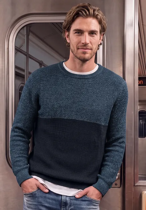 Ein Mann mit blau-schwarzem Pullover steht in einer U-Bahn, die Hände in den Taschen.