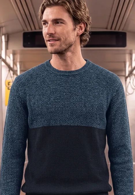 Ein Mann in einem zweifarbigen Pullover steht in einer U-Bahn oder einem Zug.