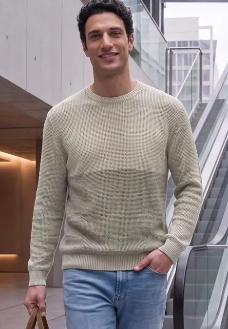 Ein Mann in zweifarbigem Pullover und Jeans steht neben Rolltreppe in modernem Gebäude.
