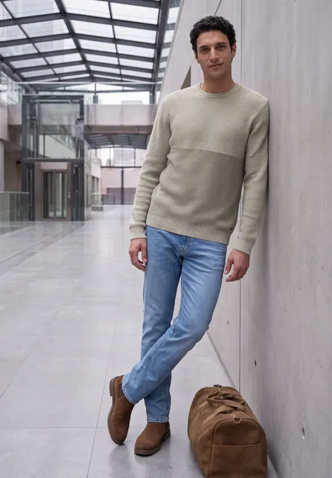 Ein lässig gekleideter Mann lehnt in einem hellen Atrium an einer Wand, neben ihm steht eine große Reisetasche.
