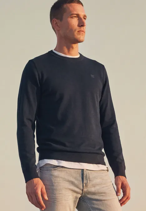 Eenvoudige trui met ronde hals in effen kleur dark basic blue Een man staat buiten en draagt een donker shirt, een wit T-shirt en een lichte spijkerbroek.