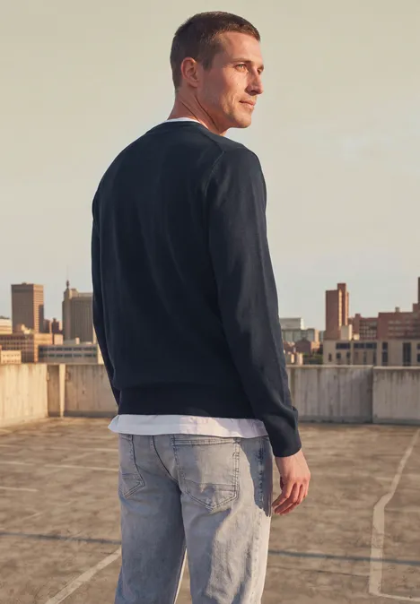 Eenvoudige trui met ronde hals in effen kleur dark basic blue Man staat op een parkeerdek, draagt een donkerblauwe trui, wit shirt en spijkerbroek, kijkt over zijn schouder.