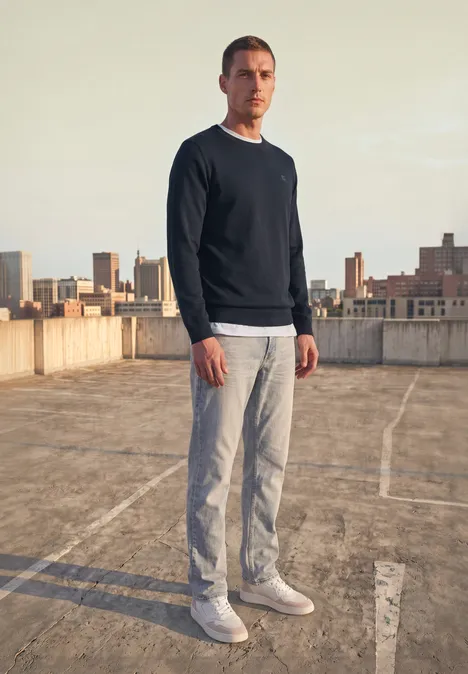 Eenvoudige trui met ronde hals in effen kleur dark basic blue Een man in trui en broek staat op een parkeerdak met de stad op de achtergrond.