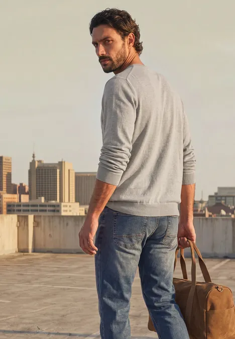 Eenvoudige trui met ronde hals in effen kleur bright beige melange Man in grijze trui en jeans staat op een dak, kijkt om zich heen en draagt een reistas.