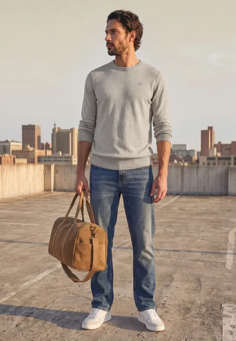 Eenvoudige trui met ronde hals in effen kleur bright beige melange Man in trui, spijkerbroek en sneakers, draagt reistas op dak met uitzicht op de stad.