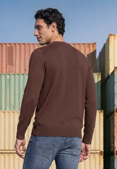 Sweter Basic z okrągłym dekoltem w jednolitym kolorze maroon brown Mężczyzna w brązowym swetrze i dżinsach stoi przed kolorowymi kontenerami, patrzy w górę.