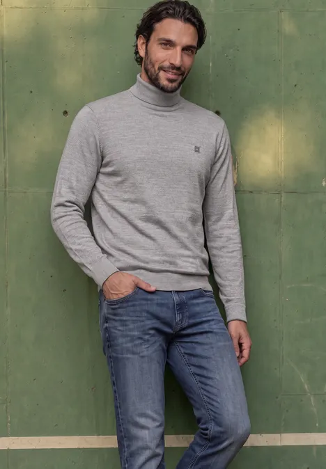 Ein Mann lehnt lässig in grauem Rollkragenpullover und Jeans vor einer grünen Wand.