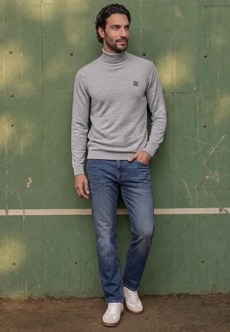 Mann mit grauem Rollkragenpullover, blauen Jeans und weißen Sneakern vor grünem Holzhintergrund.