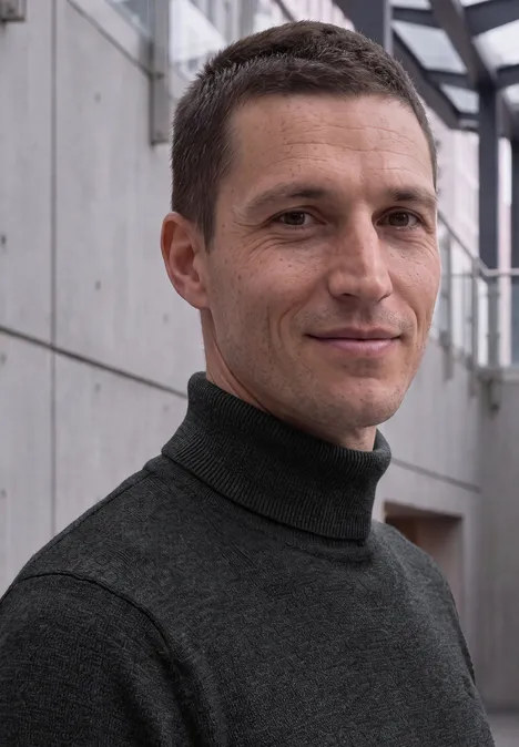 Ein Mann mit kurzen braunen Haaren in einem dunklen Rollkragenpullover steht in einem modernen Gebäude.