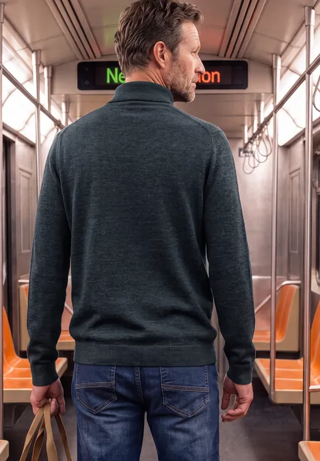 Ein Mann steht in einer U-Bahn, trägt Jeans, dunklen Pullover und hält eine Tasche, Blick nach vorn.