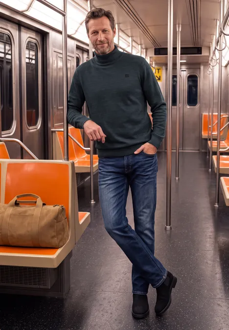 Ein Mann in Jeans und Pullover lehnt entspannt an einem Haltestab in einem fast leeren U-Bahn-Wagen mit orangen Sitzen.