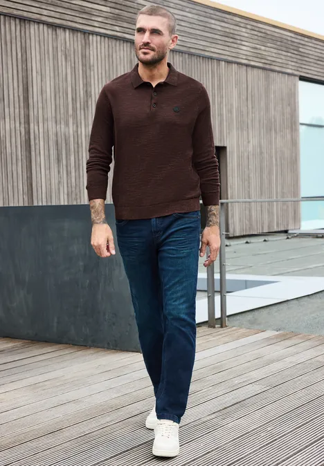 Gebreid poloshirt in mouliné-look maroon brown Een man in een bruin poloshirt, jeans en witte sneakers loopt op een houten terras voor een modern gebouw.